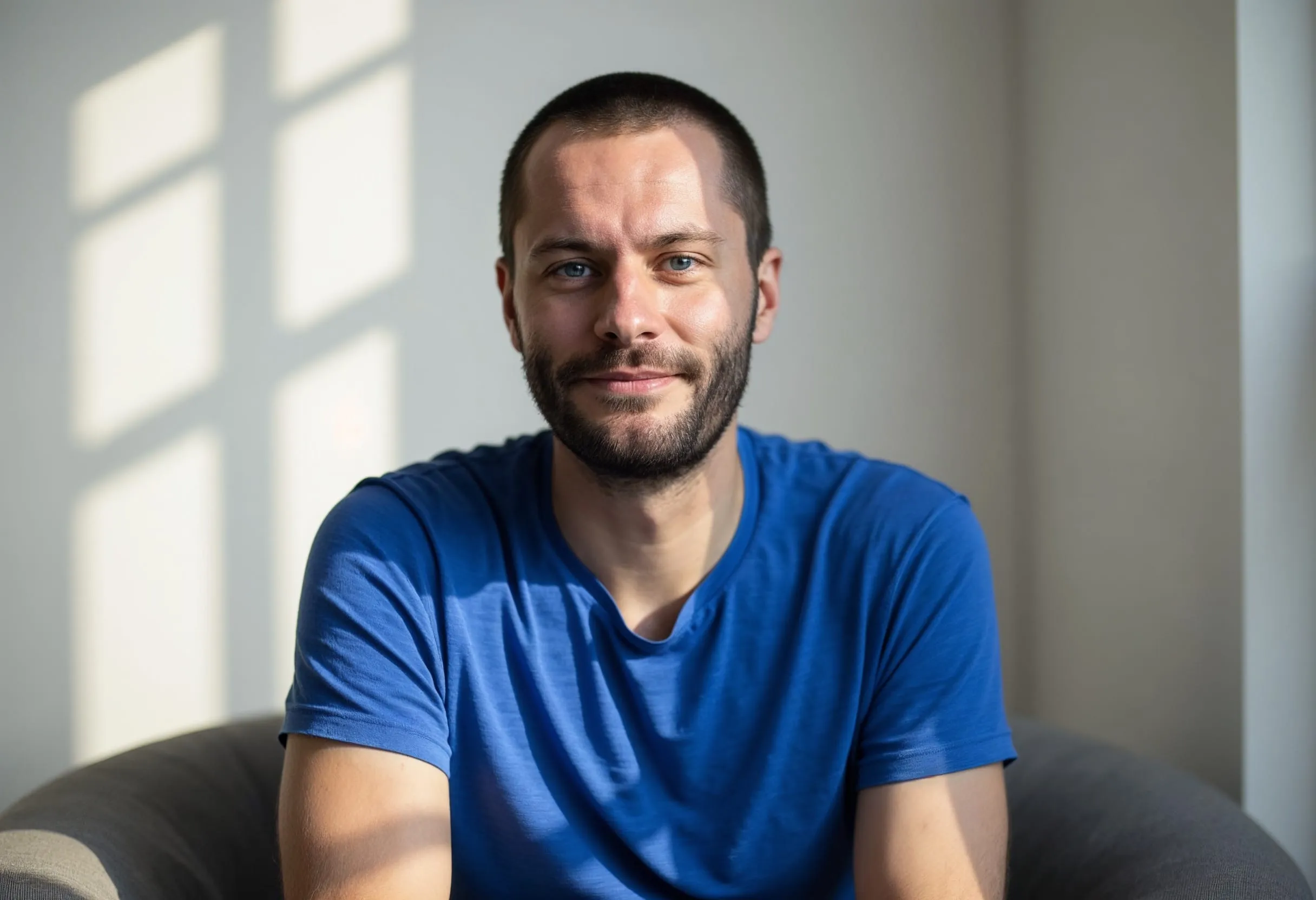 Simeon Zickert, kurze Haare und Vollbart, im blauen T-Shirt, mit leichtem Lächeln auf einem grauen Sofa sitzend